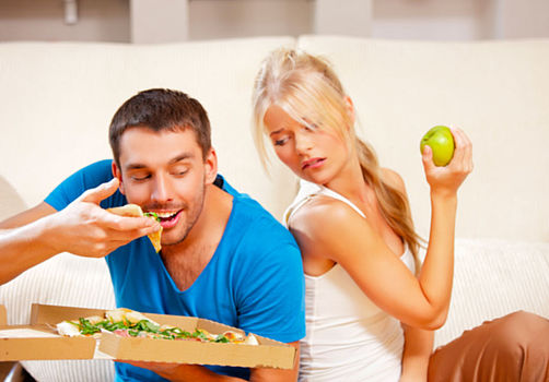 Un homme mange une pizza devant le regard envieux de sa femme, qui tient une pomme
