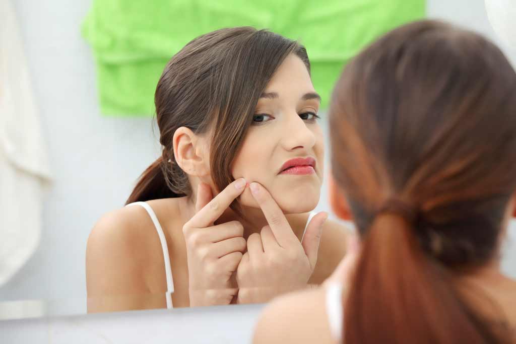 Femme éclatant un bouton d'acné devant son miroir